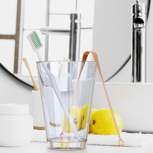 A copper tongue scraper in a cup with a toothbrush in a sink